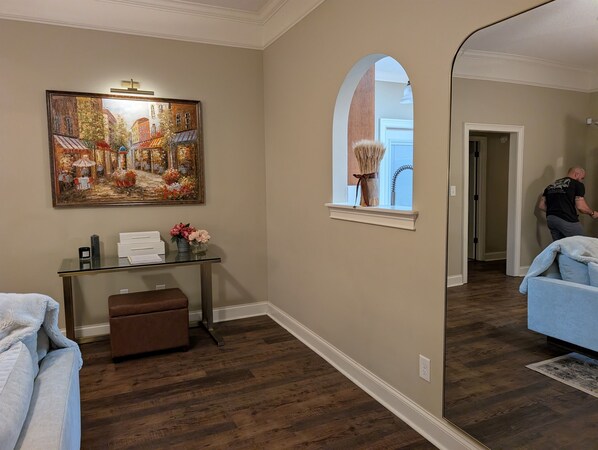 A room with a wooden floor, a glass table with a vase of flowers, a painting on the wall, and a window with a curtain.