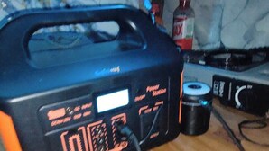 A black portable power station on a wooden surface with a bottle and a cable. - Kaixer Amsterdam (Amsterdam)