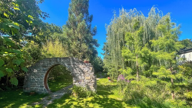 A stone archway in a lush garden with various trees and plants. - Pure relaxation: stylish nature retreat with sauna & chicken idyll (Hofheim in Unterfranken)
