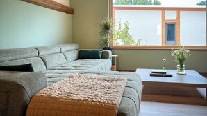 A modern living room with a grey sofa, a beige throw blanket, a wooden coffee table with a book and a vase of flowers, and a large window with a view of greenery. - Pure relaxation: stylish nature retreat with sauna & chicken idyll (Hofheim in Unterfranken)