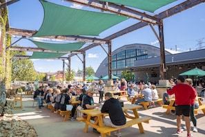 An outdoor seating area with wooden picnic tables and green canopies, surrounded by a modern building with large windows.