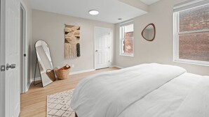A bedroom with a large bed, a mirror, a door, and a window with a brick wall view. - OTR Overview | Prime Location l Free Parking-TQL! (Cincinnati)