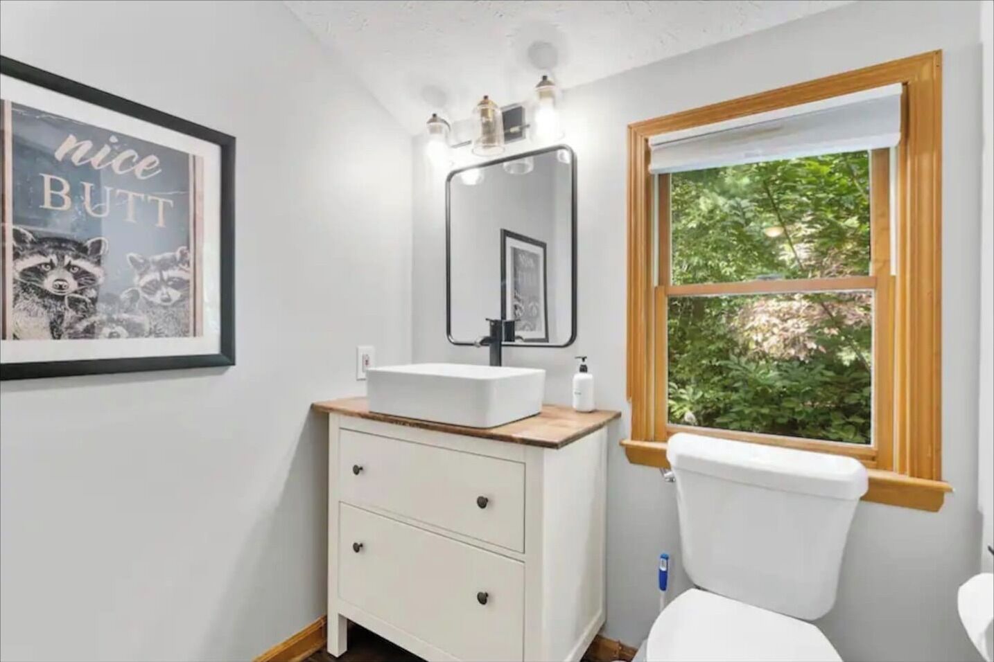 A bathroom with a white sink, a mirror, a framed picture on the wall, and a window with a view of greenery.