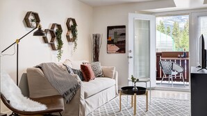 Un salon comprenant un canapé, un fauteuil, une table basse et un téléviseur. Une porte donne sur un balcon d’où l’on peut admirer la vue sur les arbres et les montagnes.