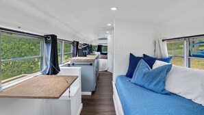 Un interior estrecho y alargado con kitchenette, comedor y una litera con ropa de cama azul.