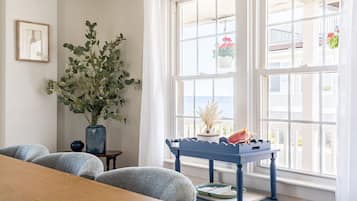 Une pièce avec une table en bois, deux chaises, une petite table d’appoint bleue agrémentée d’objets décoratifs, une grande plante et une vue sur la mer par de grandes fenêtres.