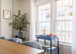 Ein Zimmer mit einem Holztisch, zwei Stühlen, einem blauen Beistelltisch mit Dekoration, einer großen Pflanze und Meerblick durch große Fenster.