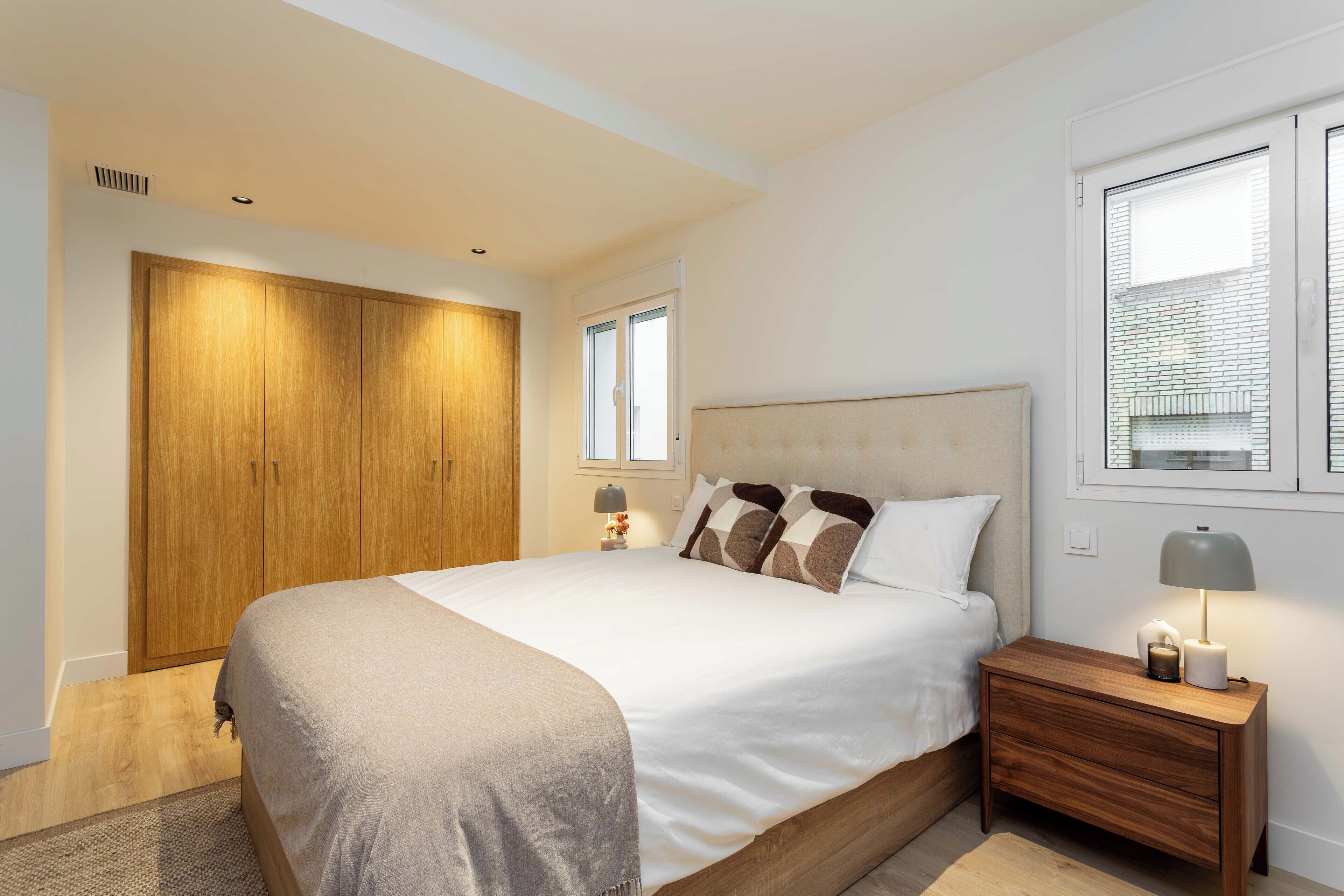 Un dormitorio con una cama grande, armario de madera, mesita de noche con lámpara y ventana con persianas.