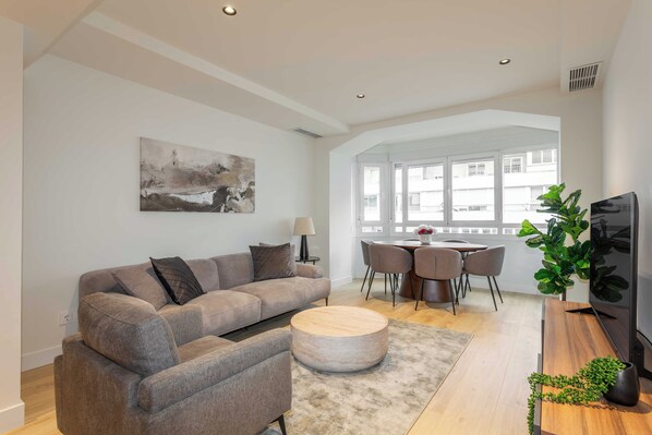 En moderne stue med en grå hjørnesofa, et rundt sofabord og et spiseområde med bord og stole.