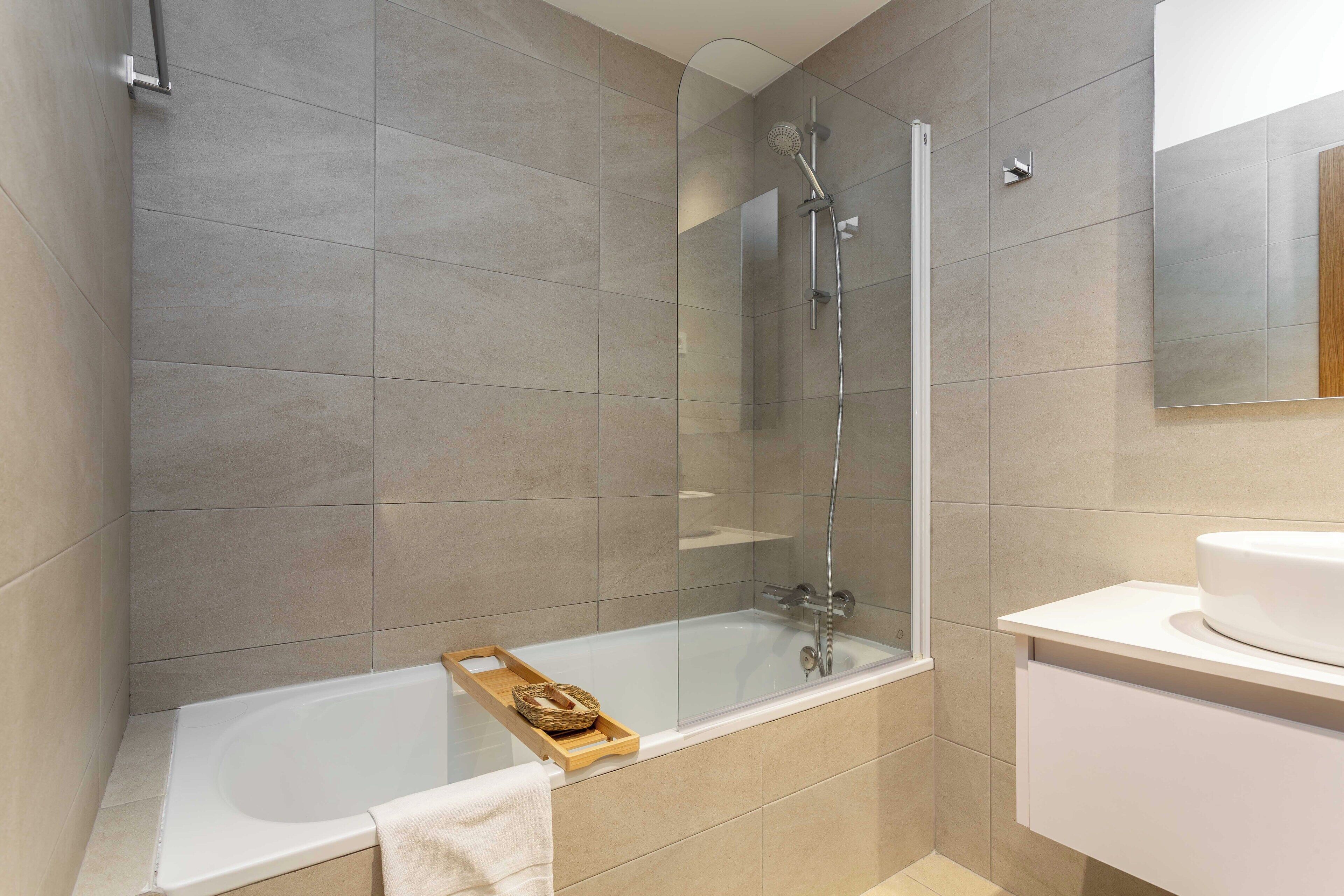 A modern bathroom with a glass-enclosed shower, a white bathtub, and a wooden tray with a basket.