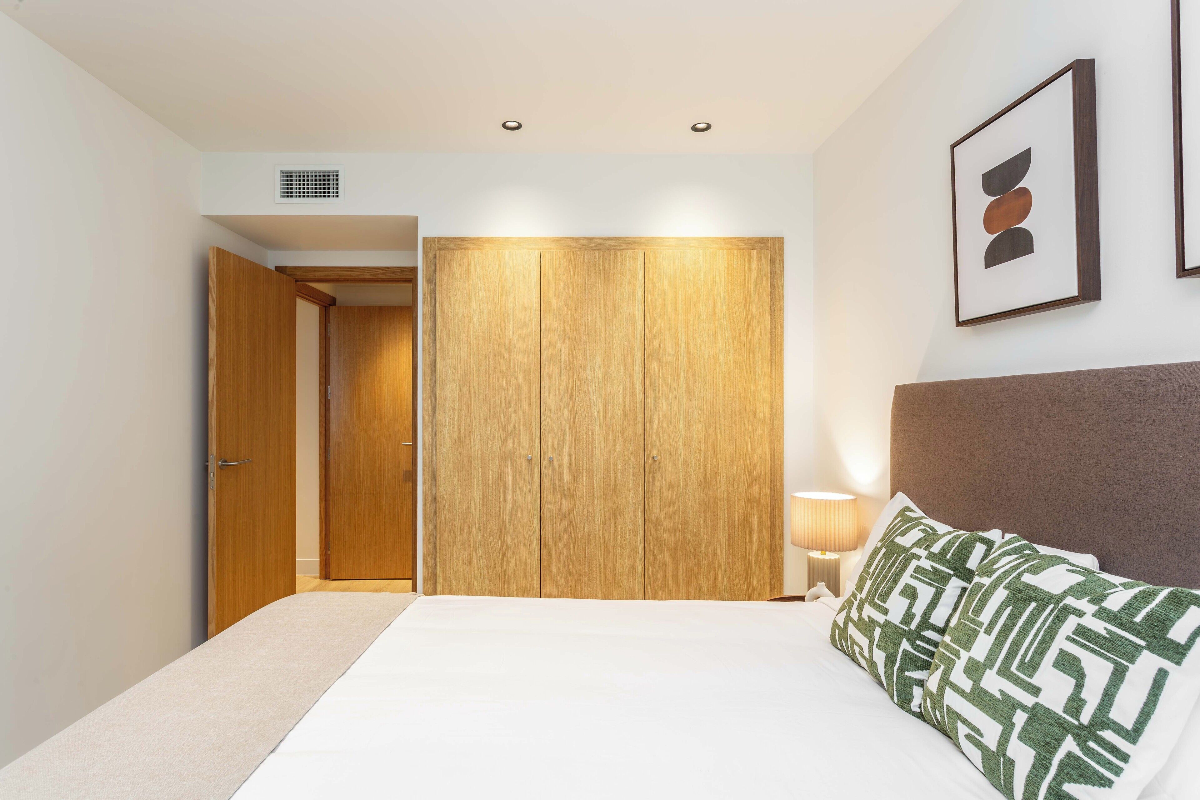 A bedroom with a bed, a nightstand, a lamp, a wooden wardrobe, and a framed picture on the wall.