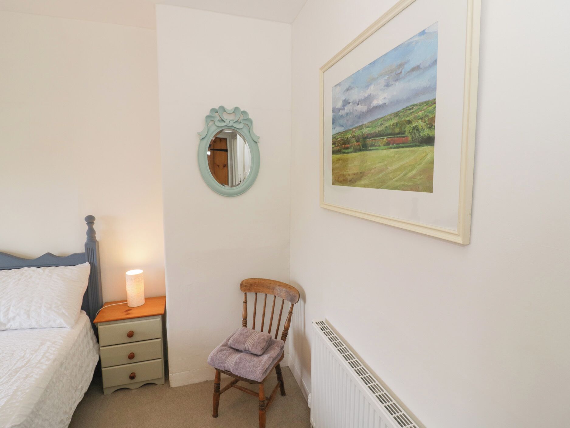 A bedroom with a bed, bedside table, lamp, chair, and a framed landscape painting on the wall.