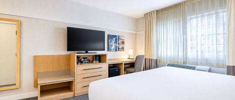 A hotel room with a large bed, a desk with a chair, a television, and a window with curtains.