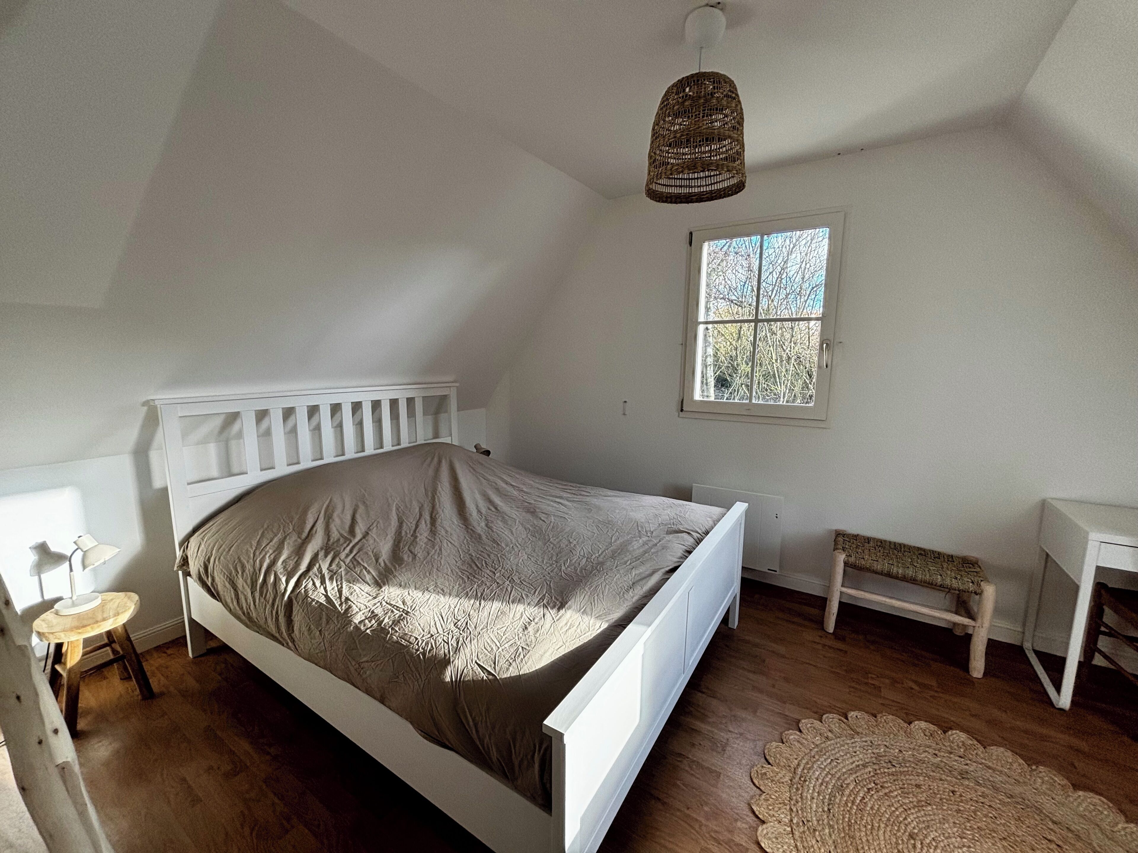 A bedroom with a white bed, a small bedside table, a window, and a woven floor lamp.