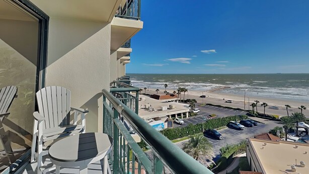 A balcony with white Adirondack chairs, a view of the beach, and a parking area. - San Luis 736 - Oceanfront Paradise (Galveston)