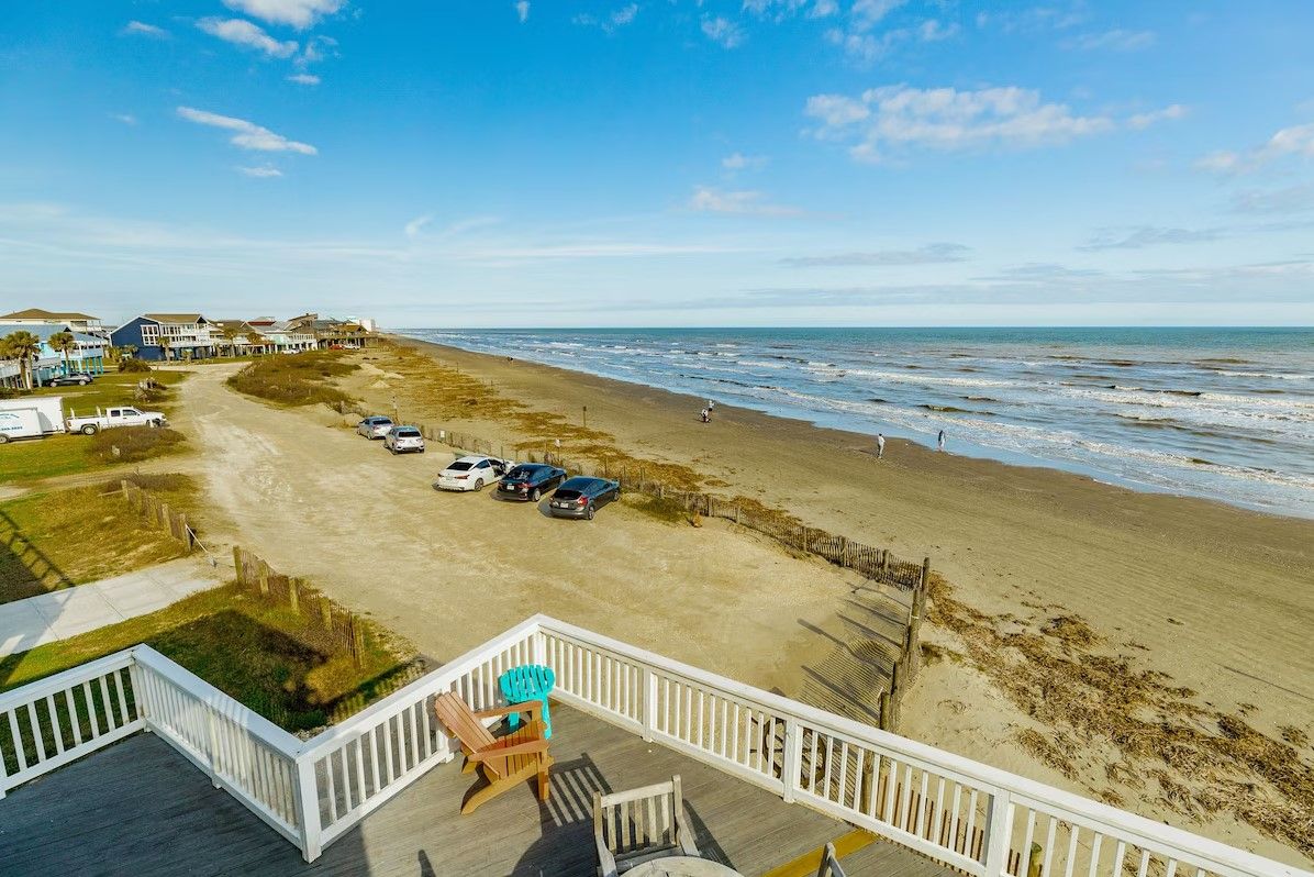 A beach view from a balcony with a deck, a parking area with cars, and a sandy beach.