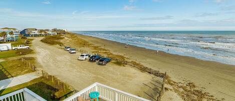 A beach view from a balcony with a deck, a parking area with cars, and a sandy beach.