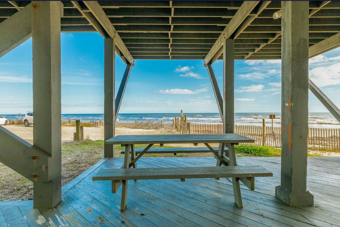 Una terraza de madera con una mesa de picnic bajo una estructura metálica, con vista a la playa y al océano.