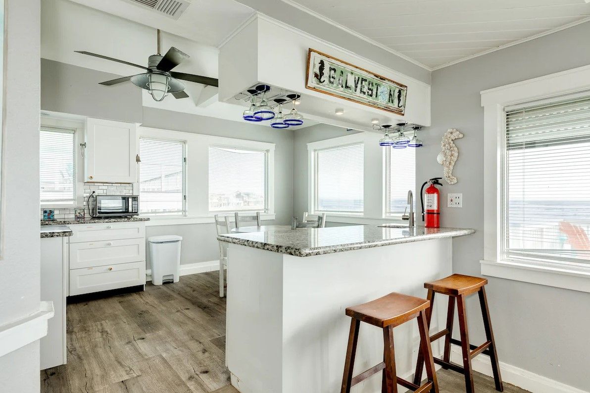 A modern kitchen with white cabinetry, a central island with stools, a ceiling fan, and a sign reading "GALVESTON."