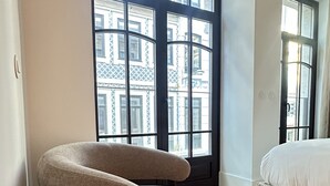Une chambre d’hôtel moderne avec un fauteuil aux lignes courbes, une petite table ronde avec une télécommande, un lit et de grandes fenêtres offrant une vue sur le paysage urbain.