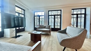 Un salon moderne doté d’un grand téléviseur à écran plat, de deux canapés, d’une table basse et de grandes fenêtres offrant une vue sur les bâtiments environnants.