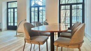 Un espace repas comprenant une table ronde et six chaises, de grandes fenêtres à encadrement noir et une vue sur les bâtiments environnants.