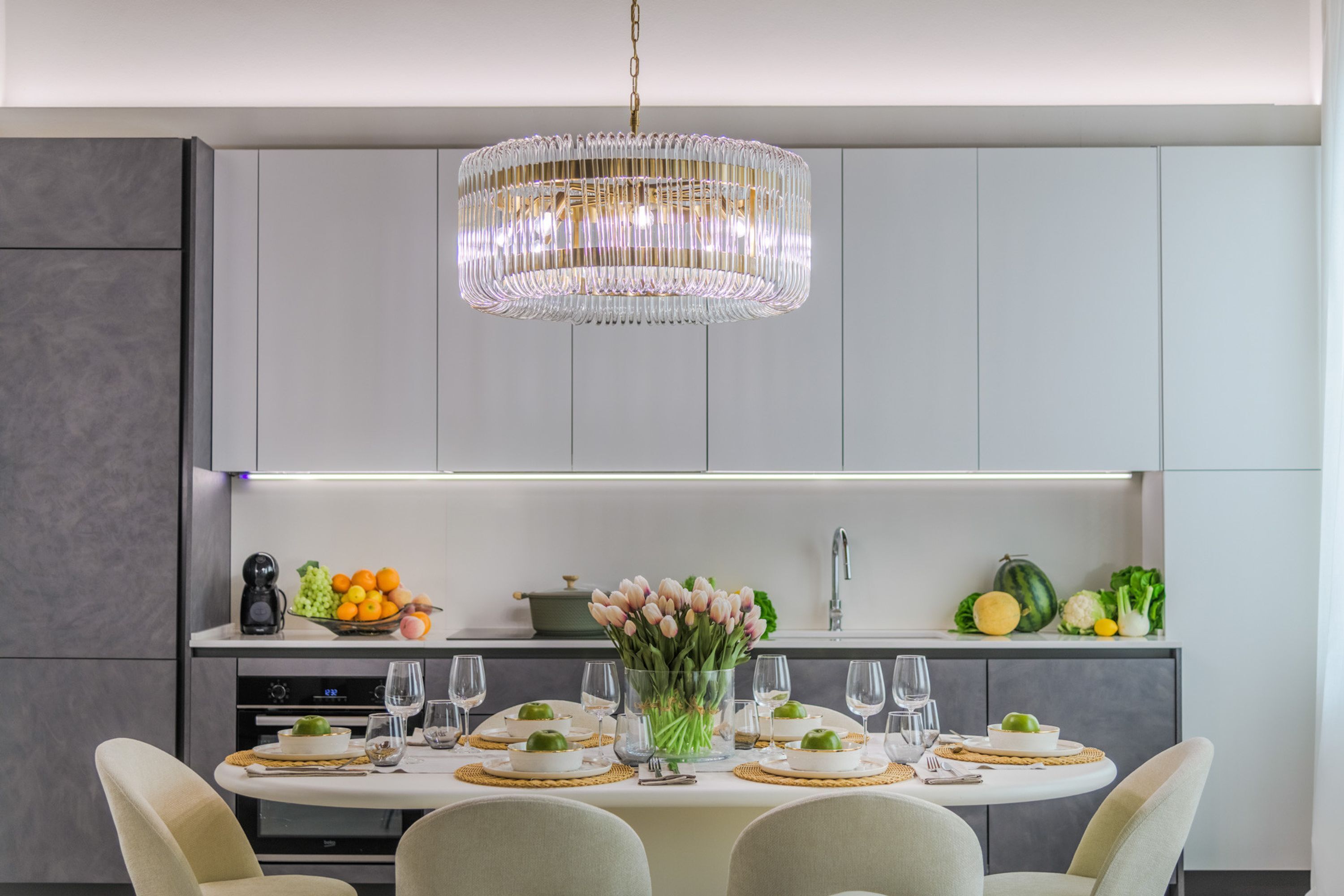 A modern kitchen with a dining table set for six, a chandelier, and a fruit bowl on the counter.