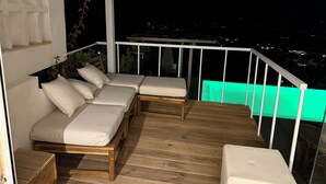 A wooden deck with a white railing, a cushioned seating area, and a view of the city at night. - Villasimius vacation home with private pool spectacular sea view golf porto (Villasimius)