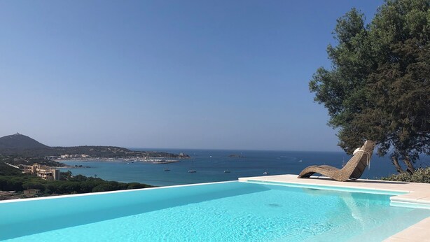 A clear blue infinity pool with a view of the sea and distant buildings. - Villasimius vacation home with private pool spectacular sea view golf porto (Villasimius)