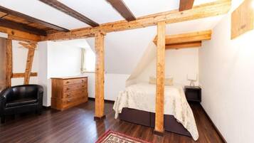 Un dormitorio con vigas de madera a la vista, una cama, un aparador, una silla y una alfombra estampada.