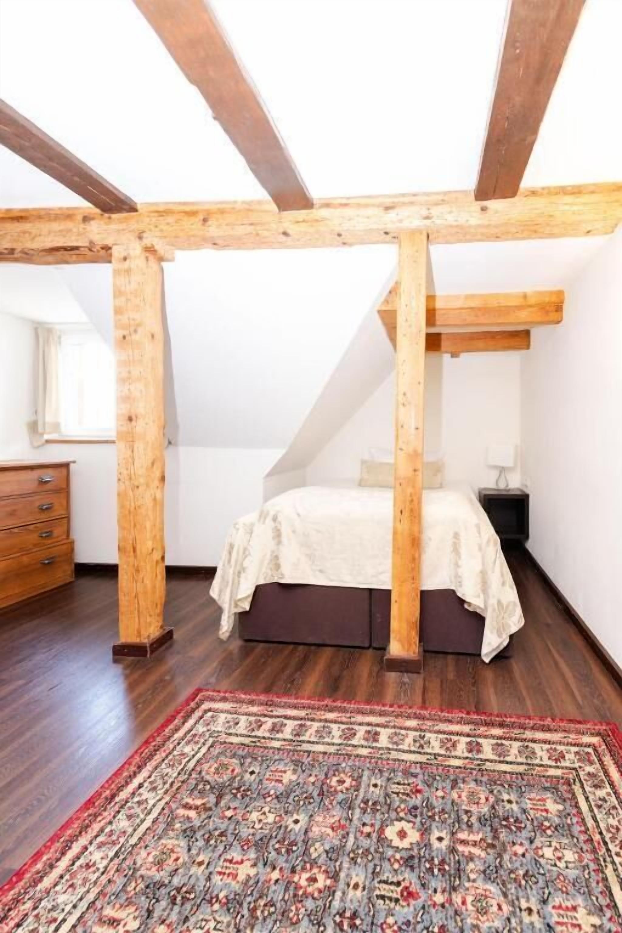 A bedroom with a sloped ceiling, wooden beams, a bed with a white bedspread, a nightstand, a dresser, and a patterned rug.