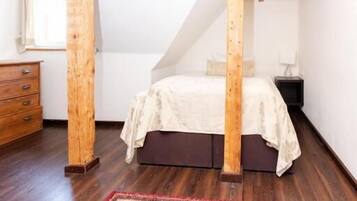A bedroom with a sloped ceiling, wooden beams, a bed with a white bedspread, a nightstand, a dresser, and a patterned rug.
