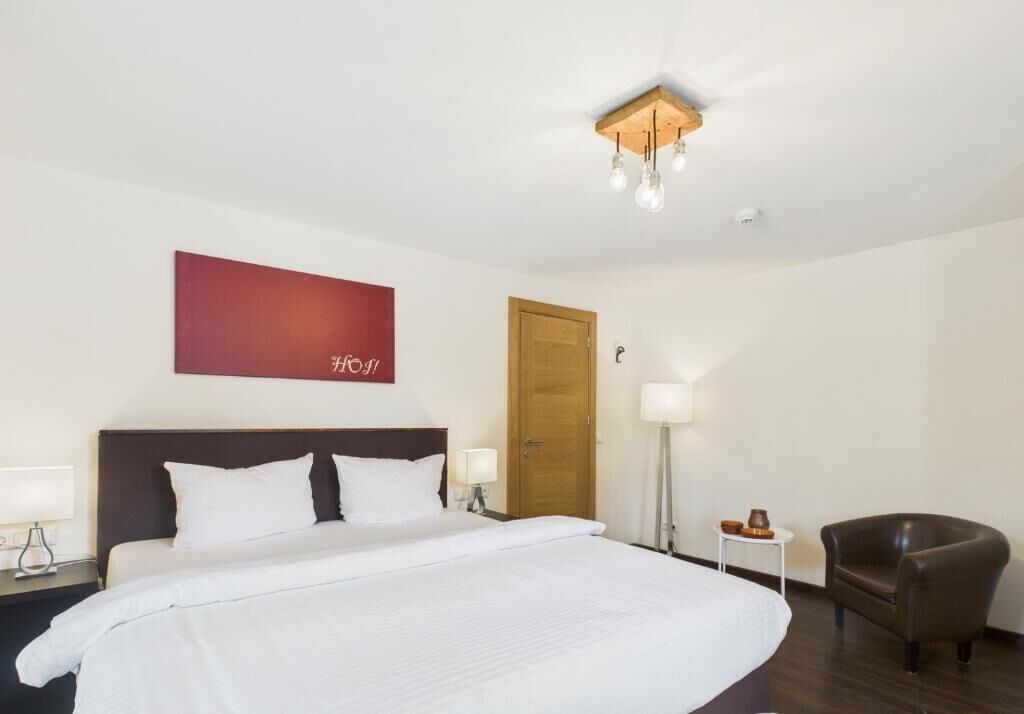 A hotel room with a large bed, a wooden door, a nightstand with a lamp, a chair, and a wall-mounted light fixture.