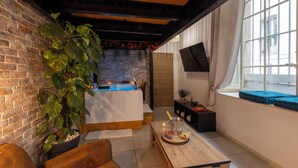 Una habitación acogedora con pared de ladrillo, una planta grande, techo de madera, televisor montado en la pared y ventana con vistas.