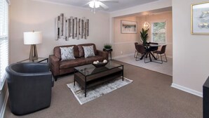 Ein Wohnzimmer mit einem braunen Ledersofa, einem Glastisch und einem schwarzen Hocker. Im Hintergrund ist eine Essecke mit Tisch und Stühlen zu sehen.
