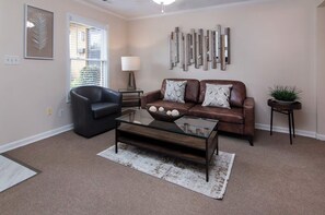 A living room with a leather sofa, a glass coffee table, a black armchair, a side table with a lamp, and a decorative wall piece. - Comfort-Ready 2BR for Work Trips (Charlotte)