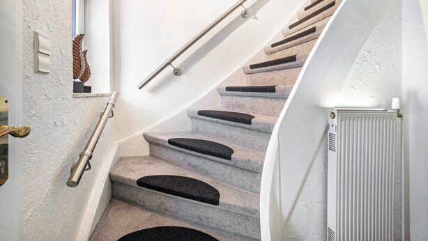 Eine Treppe mit Handlauf, Teppichstufen, einem Heizkörper und einem Fenster mit Blattdekor.