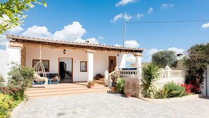 Una casa amb teulada de rajola, un porxo amb una cadira i un jardí ple de plantes diverses.