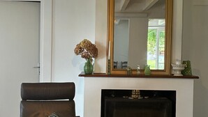 A living room with a fireplace, a large mirror, a chair, and a table. - Holiday Home 'Bienvenue À La Maison' with Private Terrace, Garden & Wi-Fi (Mézos)