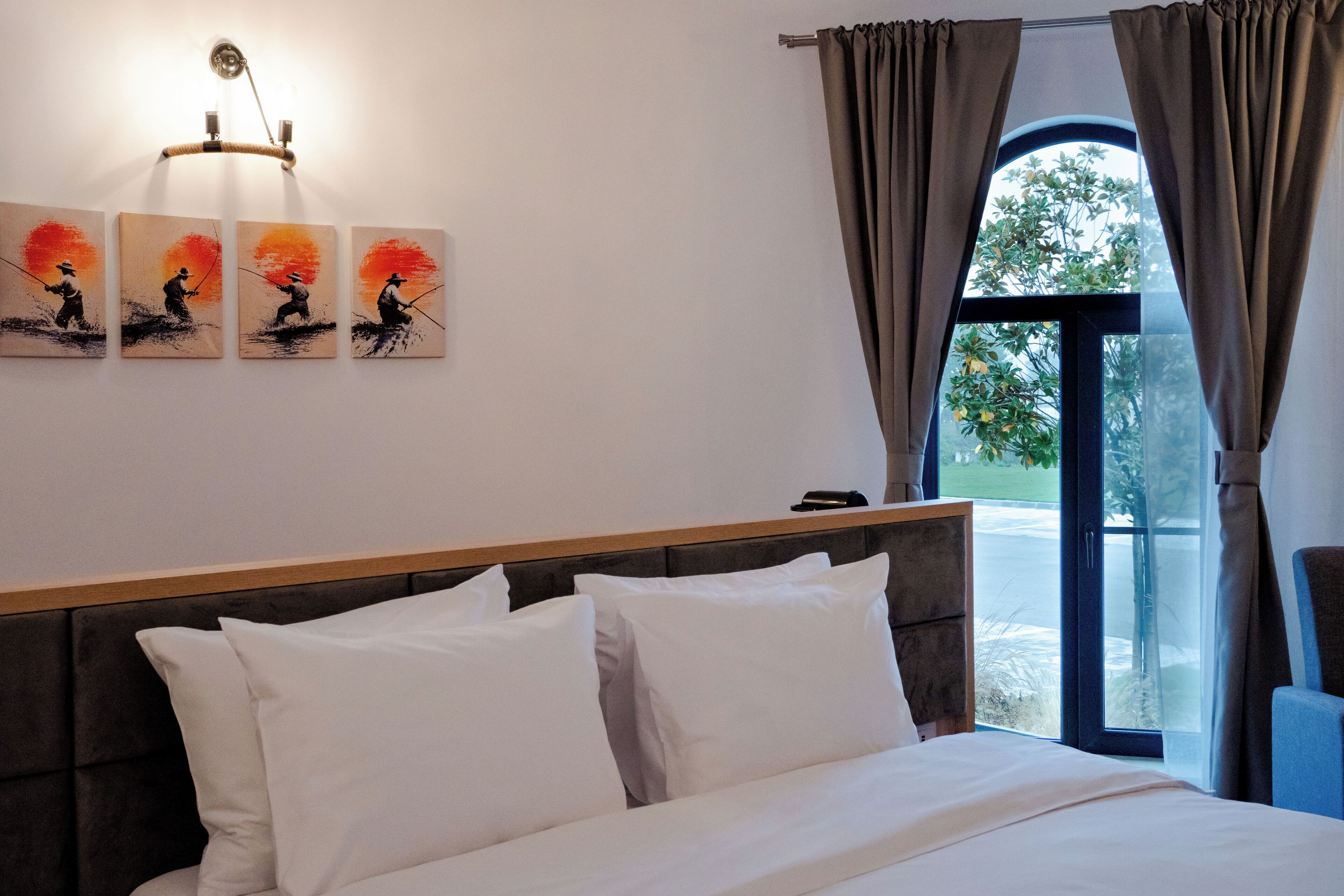 Habitación de hotel con cama, almohadillas blancas, cabecera, lámpara fijada a la pared y ventana con cortinas.