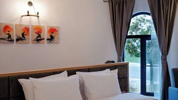 Habitación de hotel con cama, almohadillas blancas, cabecera, lámpara fijada a la pared y ventana con cortinas.