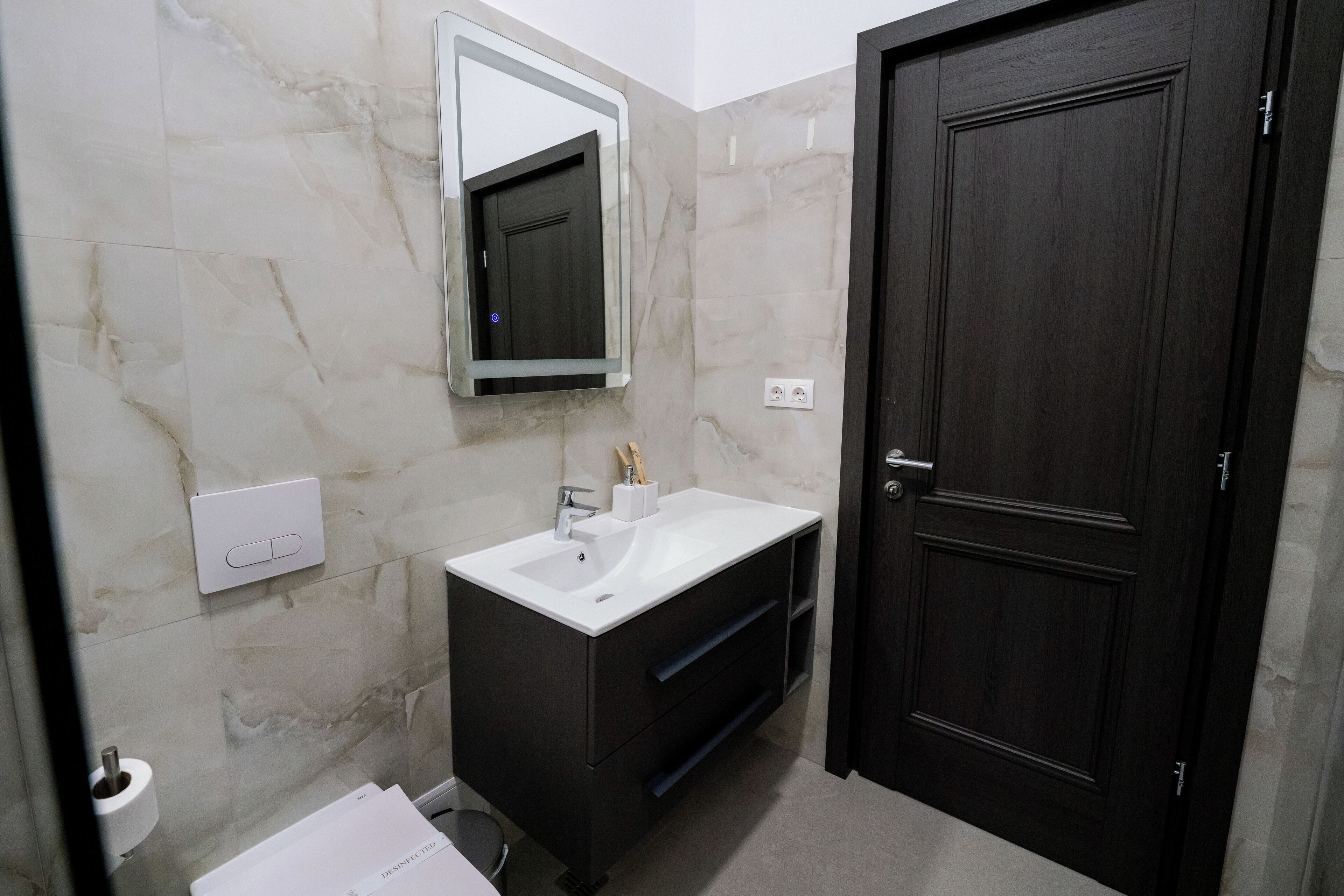 A bathroom with a marble wall, a white sink, dark cabinetry, a mirror, and a dark wooden door.