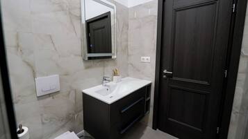 A bathroom with a marble wall, a white sink, dark cabinetry, a mirror, and a dark wooden door.