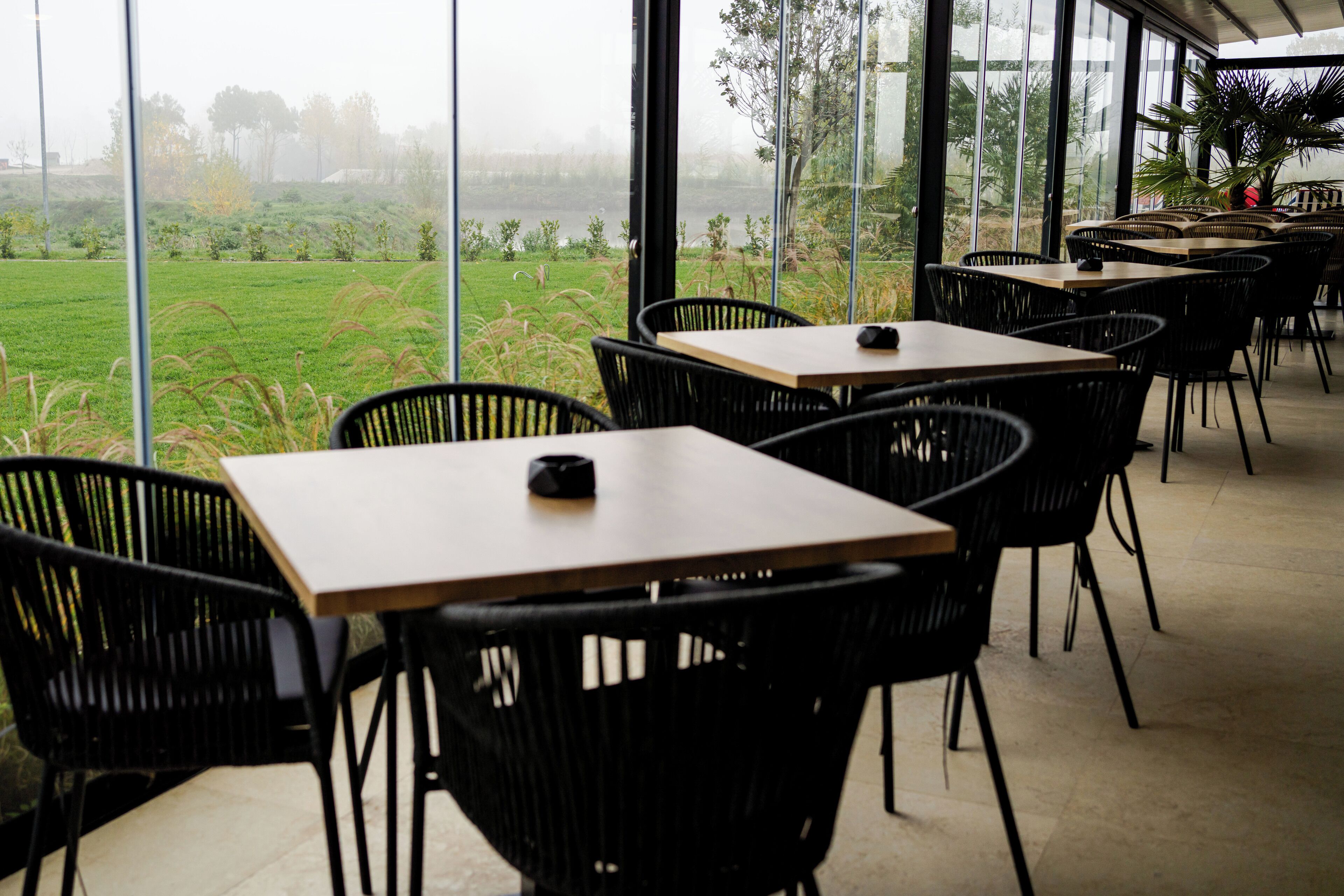 Un comedor interior con mesas y sillas, amplias ventanas que ofrecen vista a un campo cubierto de césped y árboles.