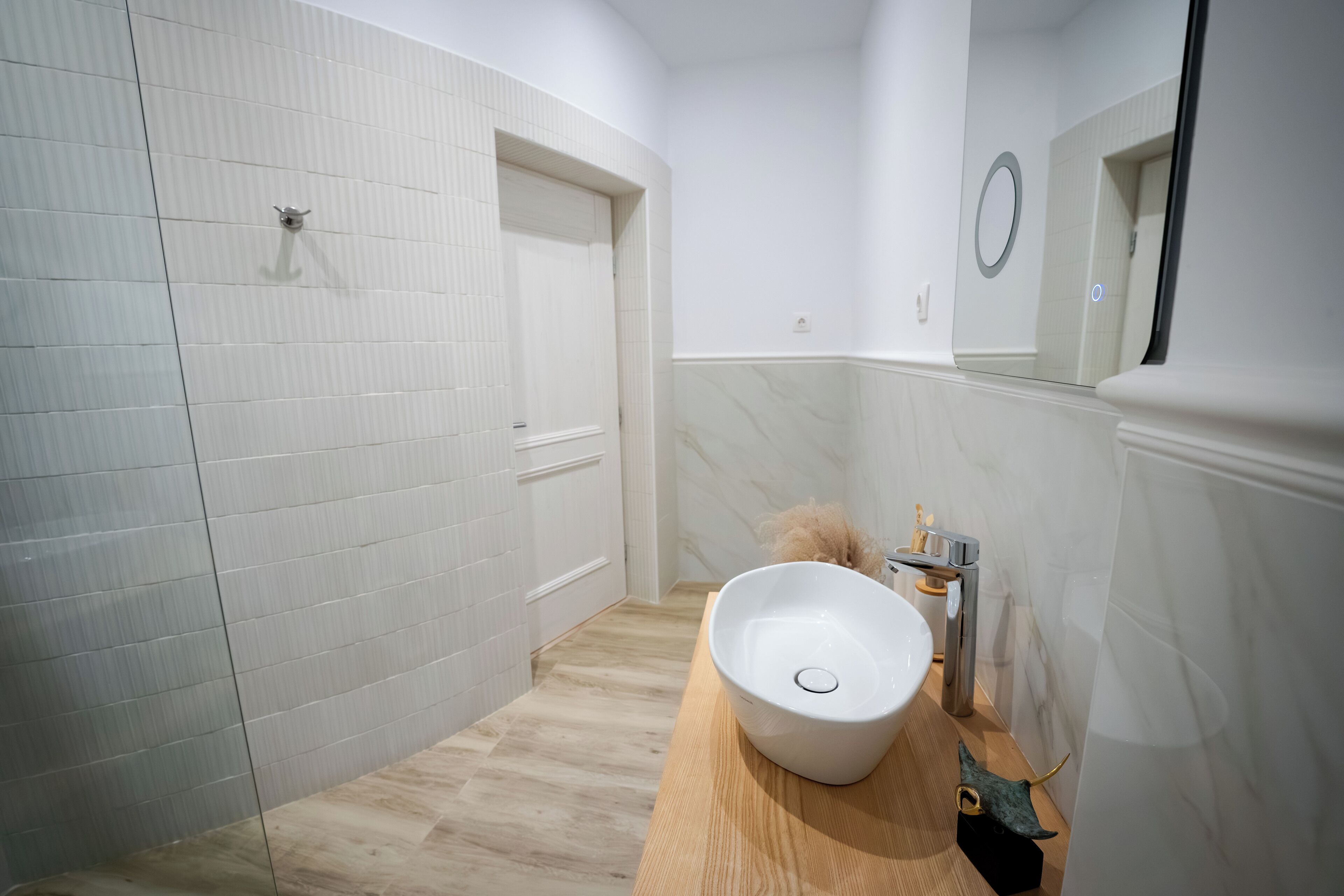 Une salle de bain avec un mur recouvert de carreaux blancs, un meuble-lavabo en bois, un lavabo blanc et un miroir.