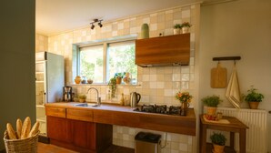 Μια κουζίνα με ένα παράθυρο, ξύλινα ντουλάπια, έναν νεροχύτη και έναν κάδο απορριμμάτων.