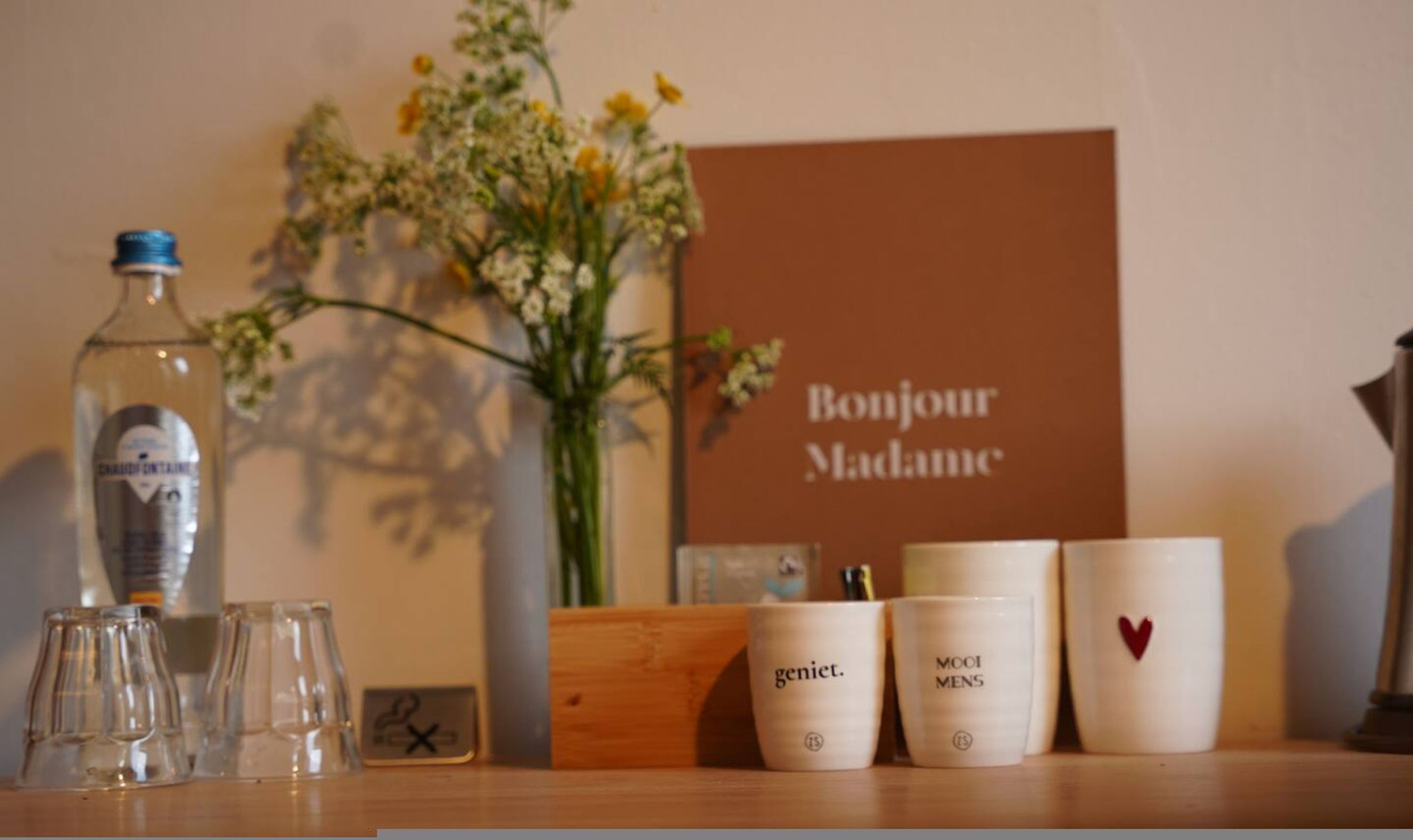 Een tafel met een fles, glazen, een vaas met bloemen en een bordje met de tekst 'Bonjour Madame'.