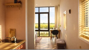 A kitchen with a stove, a dining area with a table and chairs, a window with blinds, and a view of a field. - Geau Tiny House in Weidum (Weidum)