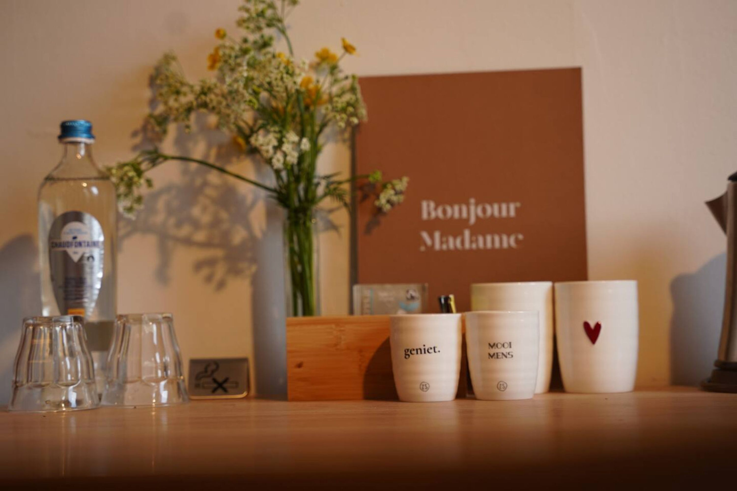 Ein Tisch mit einer Flasche, Gläsern, einer Blumenvase und einem Schild mit der Aufschrift 'Bonjour Madame'.