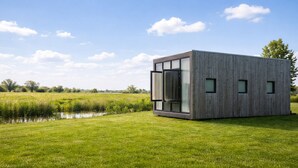 Un bâtiment moderne de forme rectangulaire, doté de grandes fenêtres en verre et d’un toit plat, situé dans un champ verdoyant, non loin d’une étendue d’eau.